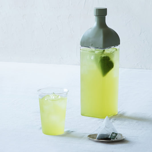 Nakamura Tokichi Sencha Tea Bag brewing in a Clear Bottle alongside iced green tea in glass cups and a fresh tea bag on a small display plate.
