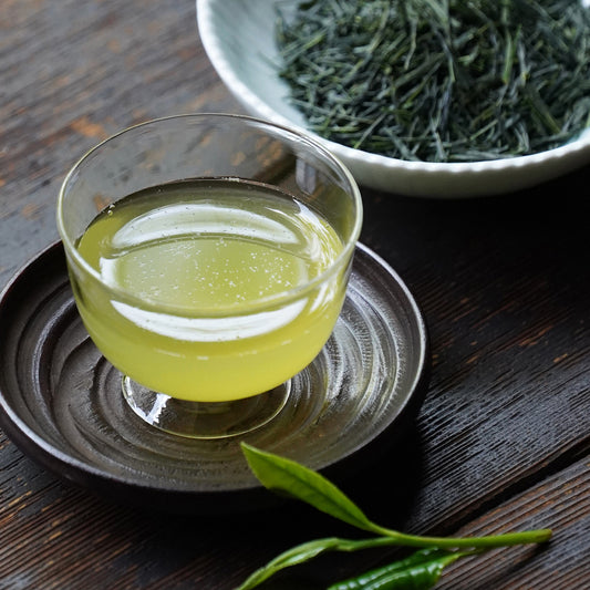 Nakamura Tokichi Shincha First Tea of the season in a glass cup on a wooden saucer, displayed with fresh loose tea leaves to highlight the seasonal Uji harvest.