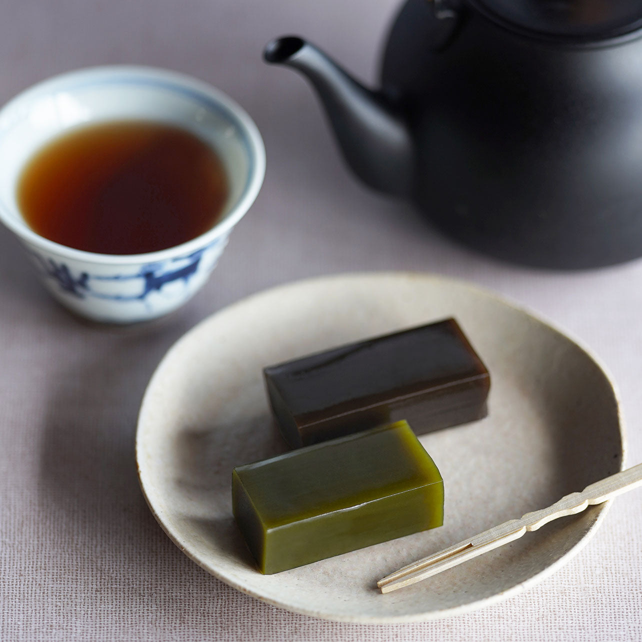 Two rectangular slices of Kogara-yokan sweet bean jelly, one deep green matcha and one reddish-brown hojicha, presented on a rustic ceramic plate with a Japanese teapot and a cup of tea in the blurred background.