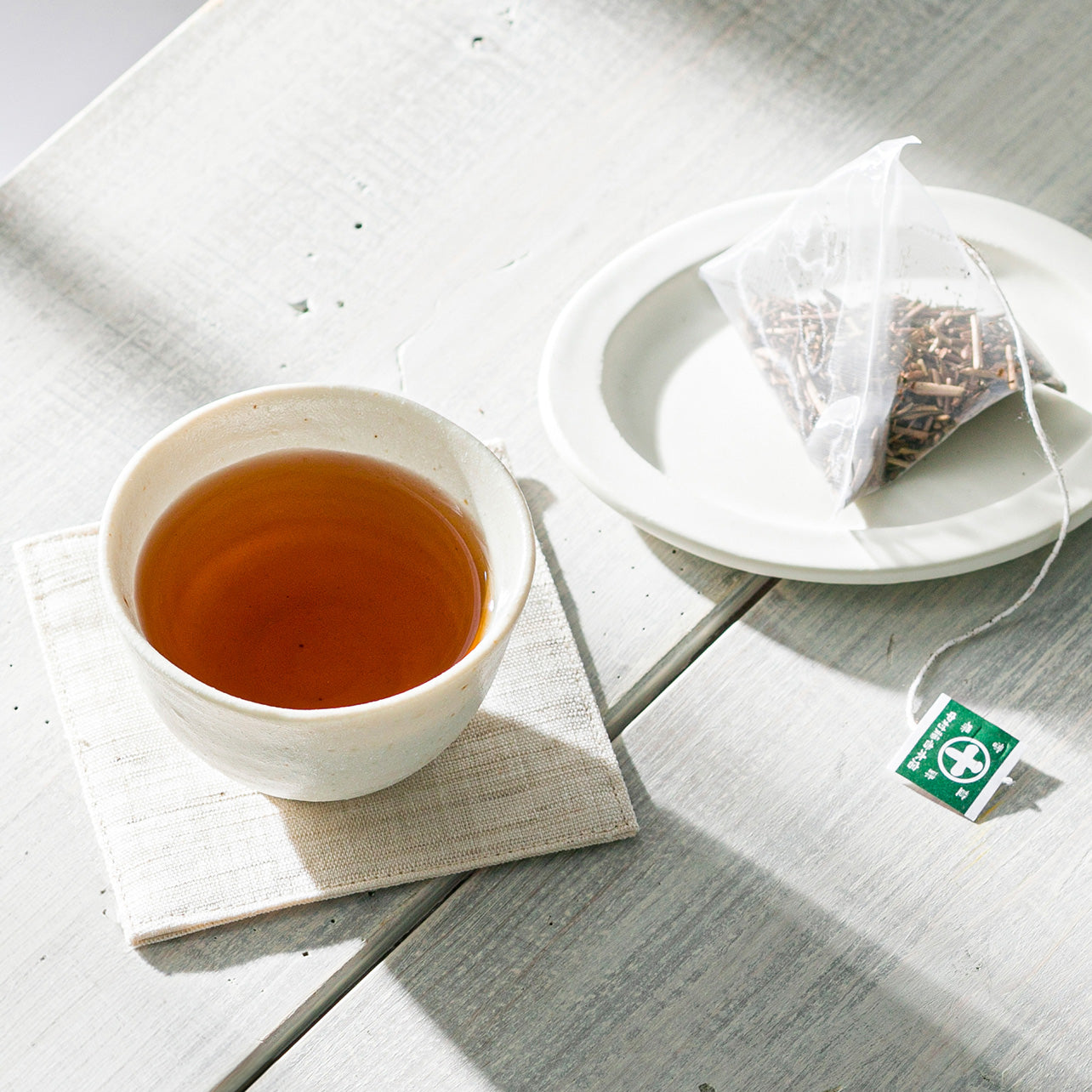 Nakamura Tokichi Hojicha Tea Bag on a small white plate next to a modern white cup of roasted green tea, set on a white wooden table in bright spring sunlight.