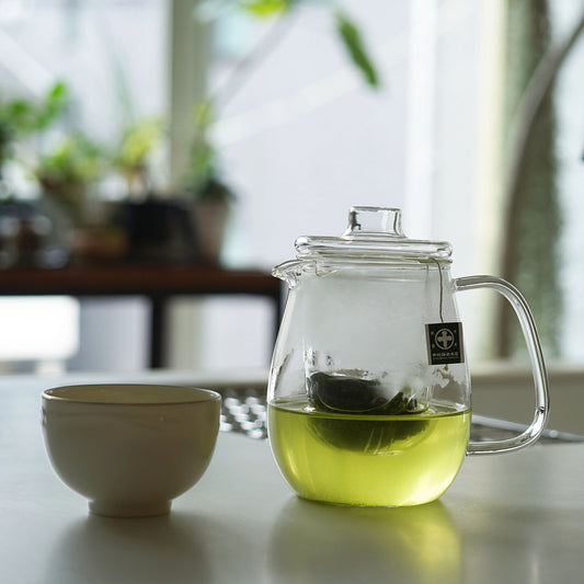 Nakamura Tokichi Japanese green tea in a clear glass teapot and cup, set against a bright, modern white room with indoor plants for a relaxing tea moment.
